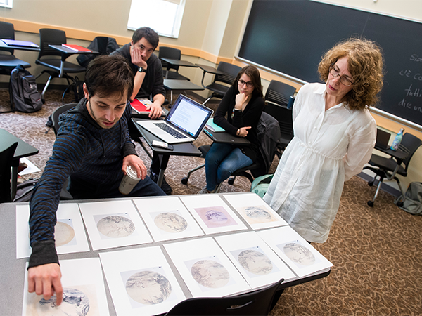 An instructor looks over a spread of prints on a desk that a student is pointing at.